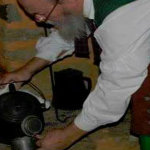 Man with colonial clothing pouring a drink from a kettle in front of a fire